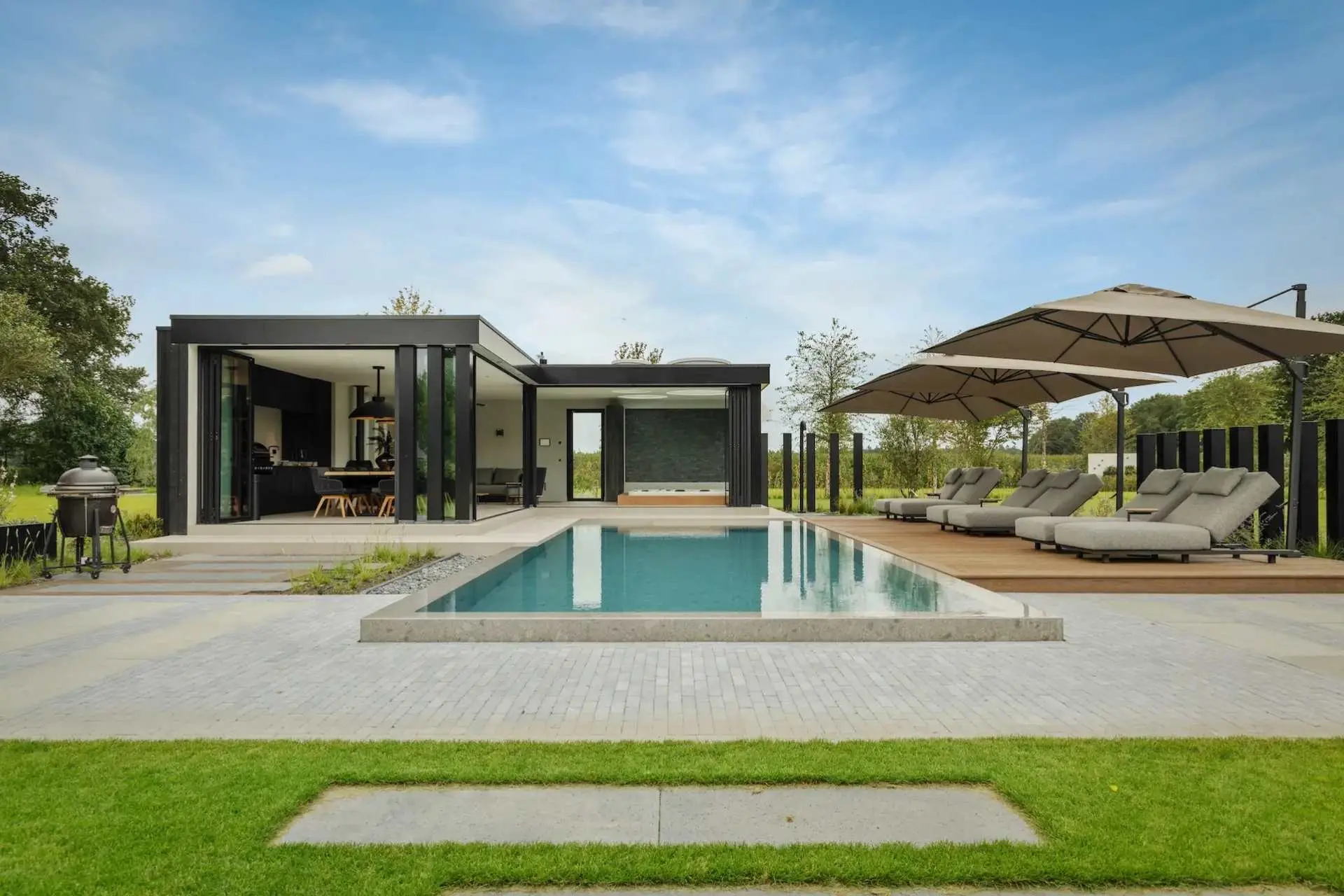 Impressie van een royaal infinity edge pool, gelegen in een riante tuin met zonneterras en modern poolhouse. Het landschapszwembad heeft natuursteenlook randen, waar het water overheen loopt.