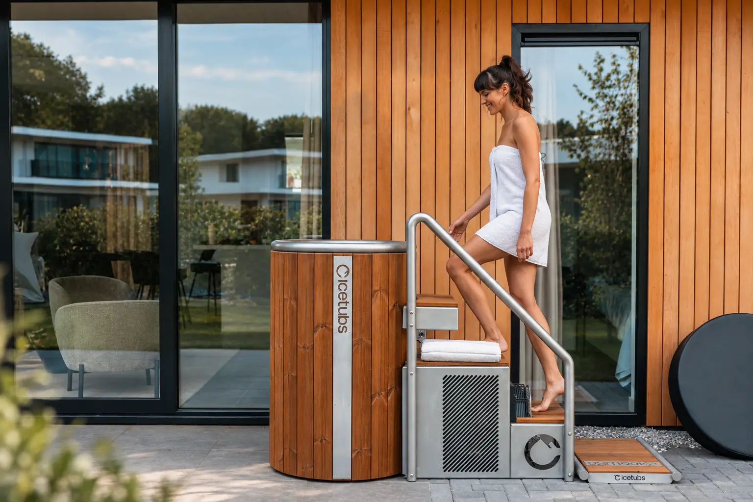 Een vrouw in een zwarte bikini loopt een modern Icetubs ijsbad met houten bekleding in, geplaatst op een terras voor een huis met houten gevel en grote glazen puien. Een rvs-koelunit met trappen en een handdoek staat naast het bad.